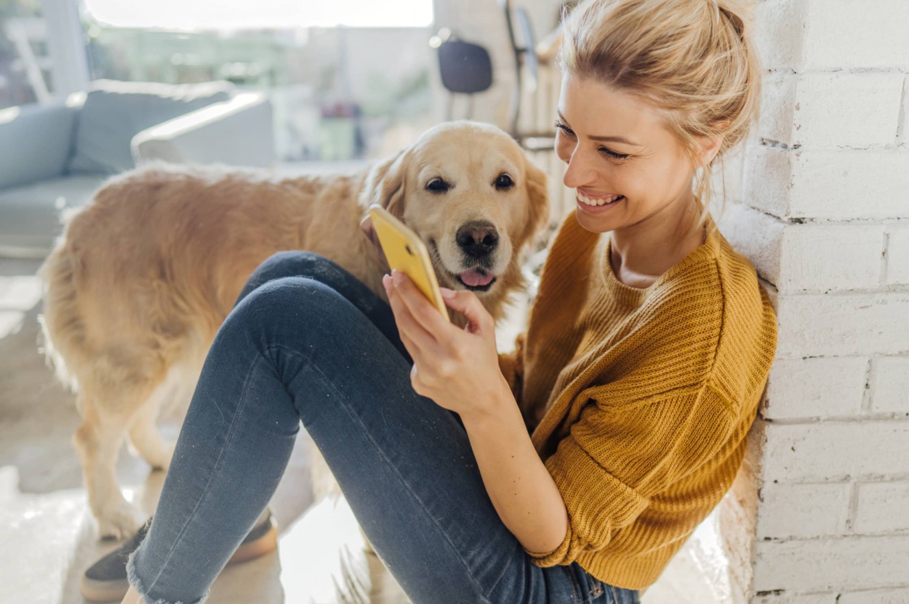 Woman in a yellow sweater smiling at her phone next to a cute yellow labrador
